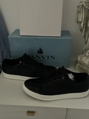 Svarta Lanvin sneakers med vit sula - Snygga svarta sneakers från Lanvin med klassisk låg profil och vit, tjock gummisula. Skorna har svarta skosnören, metallöljetter och en stilren design i läder och mocka. Perfekta för dig som gillar minimalistisk och exklusiv stil.