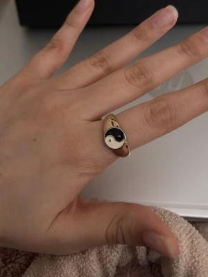 Guldfärgad ring med yin yang-symbol - Snygg guldfärgad ring med en svartvit yin yang-symbol på ovansidan. Ringen har en mjukt rundad form och symbolen är placerad centralt. Perfekt accessoar för dig som gillar unika detaljer och vill sticka ut lite extra. Anledningen till att jag säljer är pga att det är inte min stil längre. Har haft så bra minnen med den ringen. Kommer tyvärr inte ihåg vart jag köpte den ifrån