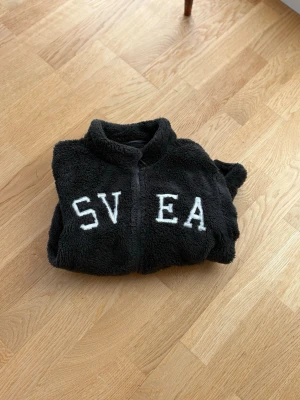 Svart teddyjacka från SVEA - Mysig svart teddyjacka från SVEA med dragkedja framtill och stora vita bokstäver på bröstet. Jackan har två fickor och en patch med SVEA-logga på ena ärmen. Perfekt för kalla dagar när du vill vara både varm och snygg.