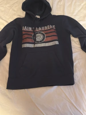  Saint Laurent hoodie - Säljer en svart hoodie från Saint Laurent med tryck i vitt och rött framtill. Hoodien har en stor magficka och klassisk huva med dragsko. Materialet känns mjukt och skönt, perfekt för en avslappnad och cool stil. Märkeslappen sitter i nacken.