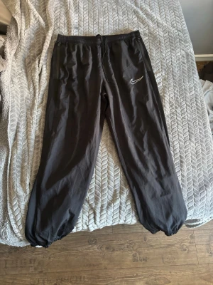 Svarta mjukisbyxor från Nike - Säljer ett par svarta mjukisbyxor från Nike med vit logga på vänster ben. Byxorna har resår i midjan med dragsko, dragkedjefickor och resår vid bensluten. Perfekta för chill eller träning, riktigt sköna och stilrena.
