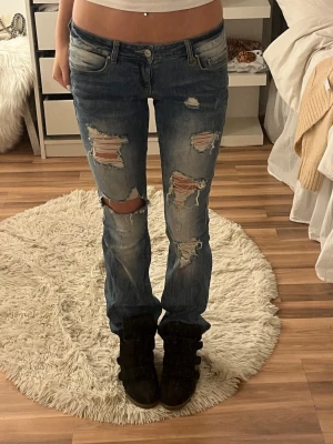 Blå slitna jeans med hål - Säljer ett par blå jeans med kraftiga slitningar och hål på både lår och knän. Jeansen har låg midja, klassisk femficksmodell och raka ben. Perfekta för en avslappnad och trendig look.