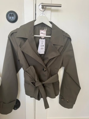 Olivgrön kort trenchcoat med skärp - Säljer en helt ny jätte fin trenchcoat från Gina Tricot i storlek xs, den är också slutsåld. Jackan har dubbelknäppning och knytskärp i midjan. Pris kan diskuteras💕💕