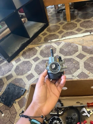 Förgasare till moped eller scooter - Säljer en metallisk förgasare som passar till moped eller scooter. Den är silverfärgad med svarta och mässingsfärgade detaljer. Kompakt och rund form med flera anslutningar för slangar och skruvar. Perfekt för dig som gillar att meka med motorfordon.