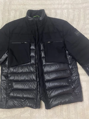 Hugo boss Jacka - Snygg svart dunjacka från BOSS med hög krage och två bröstfickor med dragkedja. Jackan har en mix av matt och glansigt material, quiltade partier och diskret BOSS-logga på ärmen. Perfekt för kalla vinterdagar och ger en modern look.