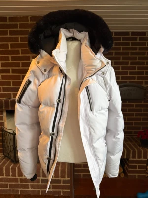 Vit Moose Knuckles dunjacka - Vit dunjacka från Moose Knuckles med svart fuskpäls på huvan och svarta dragkedjor på bröstet och ärmen. Jackan har broderad patch på insidan med hockeymotiv och texten 'Down Filled'. Perfekt för kalla vinterdagar och riktigt snygg streetstil.