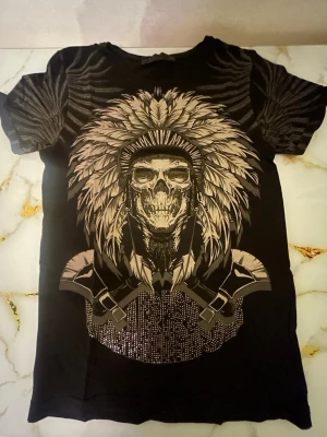 Svart t-shirt med fjädertryck - Cool svart t-shirt med stort tryck av fjädrar och detaljer framtill. T-shirten har korta ärmar och rund hals. Materialet känns mjukt och bekvämt, troligen bomull eller bomullsblandning. Perfekt för dig som vill sticka ut med en unik design.