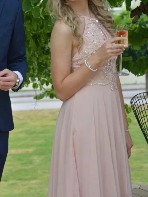 Ljusrosa långklänning med pärlor - Elegant ljusrosa långklänning med pärlbroderier och glittriga detaljer upptill. Klänningen har ärmlös design och hög halsringning, vilket ger en stilren och festlig look. Kjolen faller mjukt och materialet känns lätt och luftigt. Använd en gång. Går ned till anklarna på mig som är 173 cm lång. 