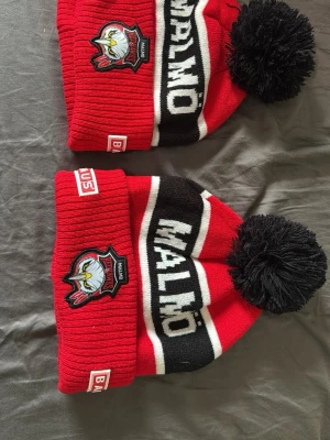 Malmö Redhawks röd mössa med tofs - Säljer två  röda och svarta Malmö Redhawks mössor med stor svart tofs på toppen. Mössorna har broderad klubbmärke framtill, vit text 'MALMÖ' och Bauhaus-logga på kanten. Perfekt för kalla dagar.