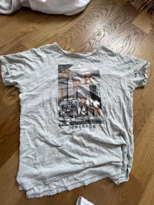 Grå t-shirt med Brooklyn-tryck - Säljer en grå t-shirt med ett stort tryck av New Yorks skyline och texten 'BROOKLYN TOMORROW' på framsidan. T-shirten har korta ärmar och en avslappnad passform. Perfekt för dig som gillar streetstyle och grafiska motiv.