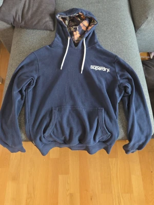 Marinblå hoodie från Superdry - Snygg marinblå hoodie från Superdry med vit logga på bröstet och vita dragsnören. Huvan har ett unikt blommigt innerfoder och magficka framtill. Perfekt för en avslappnad och trendig stil.