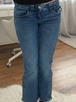 Blå bootcut jeans med broderi - Snygga blå bootcut jeans med coolt broderi och nitar på bakfickorna. Klassisk femficksmodell med knapp och dragkedja framtill. Jeansen har en lätt tvättad look och raka ben som går ut lite nedtill. Perfekta för en avslappnad och trendig stil.