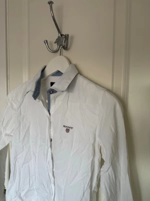 Vit skjorta från Gant med broderad logga - Klassisk vit skjorta från Gant med broderad logga på bröstet och kontrastmönstrad insida på kragen. Skjortan har långa ärmar och knappar framtill. Perfekt till både jeans och chinos för en clean look.