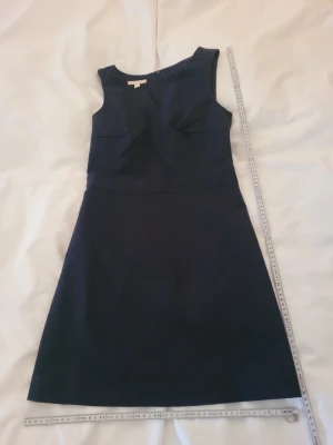 Mörkblå ärmlös klänning - Snygg mörkblå klänning med ärmlös design och lätt utsvängd kjol. Klänningen har en asymmetrisk halsringning och dragkedja i ryggen. Perfekt för dig som gillar stilrena och klassiska plagg. Materialet ser ut att vara ett fast tyg som håller formen.