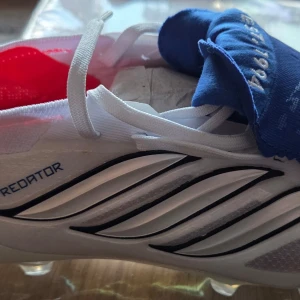 Vita Adidas Predator fotbollsskor - Snygga vita Adidas Predator fotbollsskor med svarta detaljer och klassiska vågiga linjer på sidan. Skorna har vita skosnören och röd insida. Tungan är blå med texten 'EST. 1994' broderad. Perfekta för dig som vill sticka ut på planen.