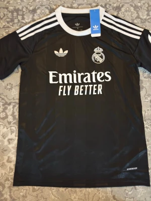 Svart Real Madrid tröja Adidas - Svart kortärmad Real Madrid tröja från Adidas med vita detaljer och klassiska tre ränder på axlarna. Tryck med Emirates Fly Better på bröstet och klubbmärke. Tillverkad i lätt och ventilerande material, perfekt för träning eller match.