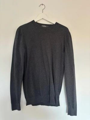 Mörkgrå stickad tröja H&M - En stilren mörkgrå stickad tröja från H&M med rund halsringning och långa ärmar. Tröjan har en enkel och klassisk design, perfekt för lager på lager och passar till det mesta. Materialet är mjukt och tunt, vilket gör den skön att bära.