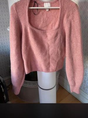Rosa stickad croppad tröja - Söt rosa stickad tröja med croppad passform och fyrkantig ringning. Tröjan har långa ärmar och ribbade muddar. Perfekt för dig som gillar en trendig och mysig look. Materialet ser ut att vara en mjuk ull- eller akrylblandning.