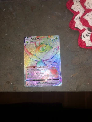 Pokémonkort Celebi VMAX Rainbow Rare - Säljer ett Pokémonkort av typen Celebi VMAX Rainbow Rare med holografisk yta. Kortet har färgglada regnbågseffekter och visar Celebi i en dynamisk pose. Perfekt för samlare eller Pokémonfans som vill utöka sin samling.