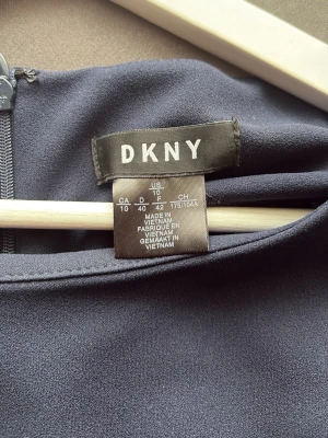 Mörkblå klänning från DKNY - Stilren mörkblå klänning från DKNY med långa ärmar och diskreta slitsar vid ärmsluten. Klänningen har en enkel, rak passform och är tillverkad i ett mjukt, följsamt material. Perfekt för dig som gillar minimalistisk och elegant stil.
