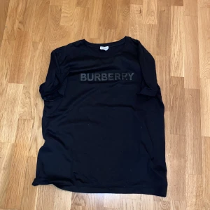Svart t-shirt från Burberry - Säljer en svart t-shirt från Burberry med stor logga i svart på bröstet. Klassisk rund halsringning och korta ärmar. Perfekt för en stilren och enkel look. Pris kan diskuteras