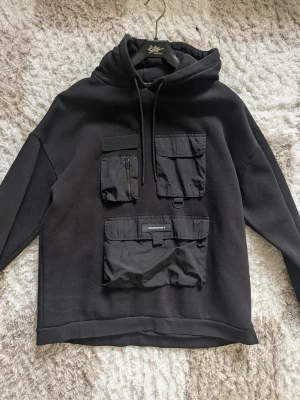 Svart hoodie med fickor och detaljer - Säljer en svart hoodie från Edgy Street med flera stora fickor framtill, dragkedja och kardborre. Hoodien har snörning i huvan och coola detaljer som metallring och patch med texten 'WARNING'. Perfekt för dig som gillar streetwear och funktionella plagg.