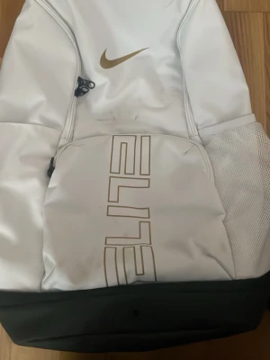 Vit Nike Elite ryggsäck - Snygg vit Nike Elite ryggsäck med guldfärgad logga och text. Ryggsäcken har flera fack, meshficka på sidan och vadderade axelband med grå detaljer. Perfekt för träning eller skola, rymmer mycket och har sportig design.