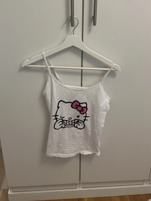 Vit Hello Kitty linnetopp från Sinsay - Säljer en söt vit linnetopp från Sinsay med smala axelband och ett tryck av Hello Kitty med rosa rosett framtill. Perfekt för sommaren och enkel att matcha med jeans eller shorts.