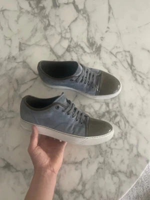 Lanvins  - Snygga sneakers i blågrå mocka med kontrasterande mörkgrå, glansig tå i lackmaterial. Skorna har vita sulor och matchande blågrå skosnören. Perfekt för dig som gillar en stilren men ändå unik look.