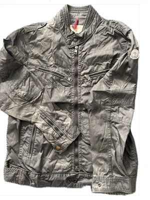 Grå vindjacka från Moncler - Snygg grå vindjacka från Moncler med hög krage, flera dragkedjeförsedda fickor och Moncler-logga på ärmen. Jackan har en sportig look, justerbara muddar och är tillverkad i ett lätt, glansigt material som står emot vind. Perfekt för dig som gillar streetwear och stilren design.