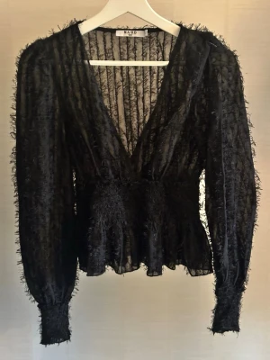Svart transparent blus från NA-KD - Snygg svart transparent blus från NA-KD med djup v-ringning och långärmad design. Blusen har ett fluffigt, strukturerat material och markerad midja som ger en cool siluett. Perfekt att styla med ett linne under för en trendig look.