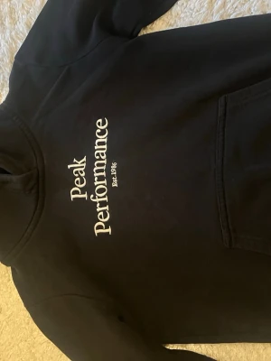 Svart hoodie Peak Performance - Säljer en svart hoodie från Peak Performance med stor vit logga på bröstet och känguruficka framtill. Klassisk modell med mjukt material och ribbade muddar. Perfekt för en avslappnad och sportig stil.