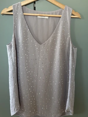 Grå paljettprydd linnetopp från Oasis - Säljer en grå ärmlös linnetopp från Oasis med v-ringning och små silverfärgade paljetter över hela framsidan och baksidan. Toppen har en rak passform och är tillverkad i ett lätt, syntetiskt material som känns luftigt. Storlek 10.