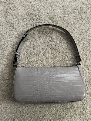 Grå axelväska - Snygg grå axelväska som är stilren och passar till allt. Väskan har ett justerbart axelband och stängs med dragkedja.