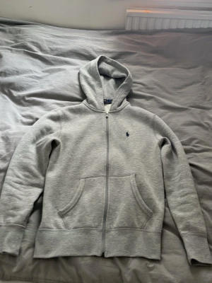 Grå hoodie från Polo Ralph Lauren - Klassisk grå hoodie från Polo Ralph Lauren med dragkedja framtill och två stora fickor. Hoodien har en liten broderad logga på bröstet och är tillverkad i mjuk bomullsmix. Perfekt för en chill och stilren look.