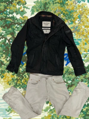 Svart feildjacket Abercrombie & Fitch - Snygg svart jacka från Abercrombie & Fitch med dragkedja och flera fickor framtill. Jackan har en klassisk krage och detaljerade sömmar som ger en cool look. Perfekt för dig som vill ha en stilren och funktionell jacka.