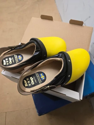 Gula och svarta trätofflor Star Clogs - Säljer ett par trätofflor från Star Clogs med gul tå och svart läderrem. Svensktillverkade med träfotbädd och justerbar rem runt hälen. Perfekta för dig som vill sticka ut med färgglada och klassiska träskor. Storlek 42 