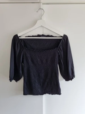 Svart smockad topp med puffärm Medicine M - Trendig svart topp med smockad kropp och puffiga trekvartsärmar. Fyrkantig ringning och elastiska ärmslut ger en snygg och bekväm passform. Perfekt till jeans eller kjol för en cool look. Fint skick