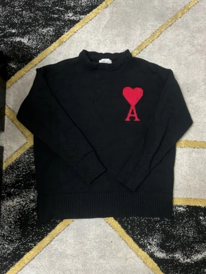 Svart stickad tröja från Ami Paris - Svart stickad tröja från Ami Paris med röd hjärtlogga och bokstaven A på bröstet. Tröjan har rund hals, ribbade muddar och är tillverkad i mjukt material. Perfekt för dig som gillar stilrena plagg med en twist.