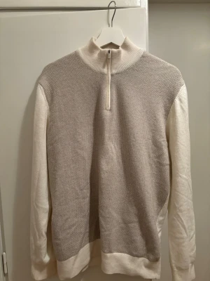 Beige stickad tröja från Boggi - Snygg beige stickad tröja från Boggi med halv dragkedja och hög krage. Tröjan har en strukturerad framsida i ljusbeige och släta vita ärmar samt muddar. Perfekt för dig som gillar stilrena och klassiska plagg i mjukt material.