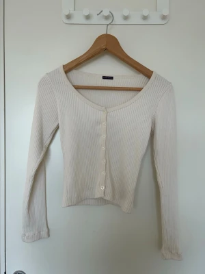 Brandy cardigan med knappar - Vit ribbad cardigan med långa ärmar och v-ringning. Tröjan har en croppad passform.