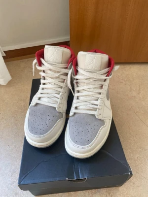 Nike Air Jordan 1 Mid grå/vit/röd - Säljer mina Nike Air Jordan 1 Mid sneakers i grått och vitt med röda detaljer. Skorna har mockaliknande material på tå och sidor, klassisk swoosh och Air Jordan-logga på sidan. Röd vadderad krage och vita skosnören. Perfekta för dig som gillar streetwear. Priset kan diskuteras vid snabb affär!!