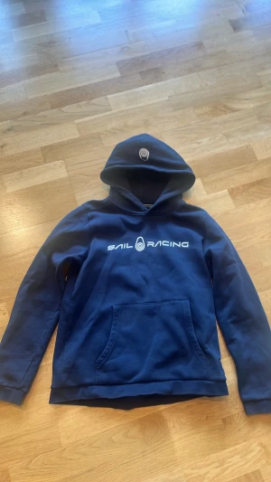 Blå hoodie från Sail Racing - Mörkblå hoodie från Sail Racing med vit logga på bröstet och tryck på huvan. Klassisk känguruficka framtill och mjukt material som känns skönt mot huden. Perfekt för dig som gillar en sportig och clean stil.