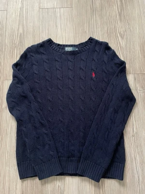 Mörkblå kabelstickad tröja Polo Ralph Lauren - Kabelstickad mörkblå tröja från Polo Ralph Lauren med rund halsringning och röd broderad logga på bröstet. Tröjan har långa ärmar och ribbade muddar vid ärmslut och nederkant. Perfekt för dig som gillar klassisk och stilren look.