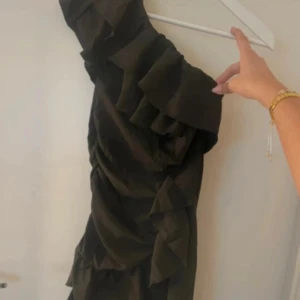 Svart omlottklänning med volanger - Snygg svart kortklänning med omlott-design och volangdetaljer längs hela klänningen. Klänningen har oneshoulder och är tillverkad i ett lätt, mjukt material som ger en elegant känsla. Perfekt för dig som vill sticka ut med en trendig look.