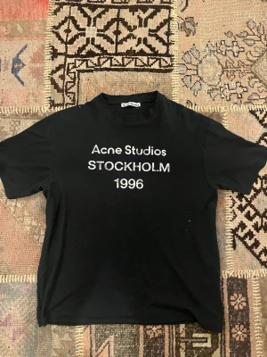 Svart Acne Studios t-shirt - Säljer en svart t-shirt från Acne Studios med vit text 'STOCKHOLM 1996' på bröstet. Klassisk rund hals och korta ärmar. T-shirten har en normal passform och är tillverkad i mjuk bomull, perfekt för en clean och stilren look.