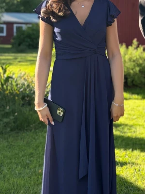 balklänning  - Elegant marinblå långklänning med omlottliv och v-ringning. Klänningen har korta volangärmar och ett mjukt, flytande tyg som ger ett snyggt fall. Perfekt för speciella tillfällen och stilrena outfits.