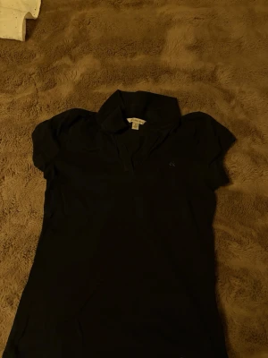 Svart pikétopp från Calvin Klein - Snygg svart pikétopp från Calvin Klein med liten 'ck'-logga broderad på bröstet. Klassisk krage och korta ärmar ger en clean och tidlös look. Perfekt till jeans eller kjol för en enkel men stilsäker outfit. Knappt använd