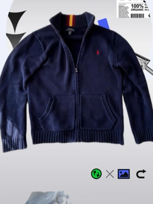 Mörkblå stickad ziptröja Polo Ralph Lauren - Mörkblå stickad ziptröja från Polo Ralph Lauren med röd broderad logga på bröstet. Tröjan har ribbad krage med röd och gul rand, två fickor fram och ribbade muddar. Perfekt för lager på lager och snygg till jeans.