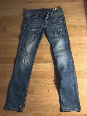 Blå slitna jeans med patchdetalj - Säljer ett par blå jeans med slitningar och patchdetalj på höger lår. Jeansen har klassisk femficksmodell, raka ben och en cool tvättad look. Perfekta för dig som gillar en avslappnad och trendig stil.